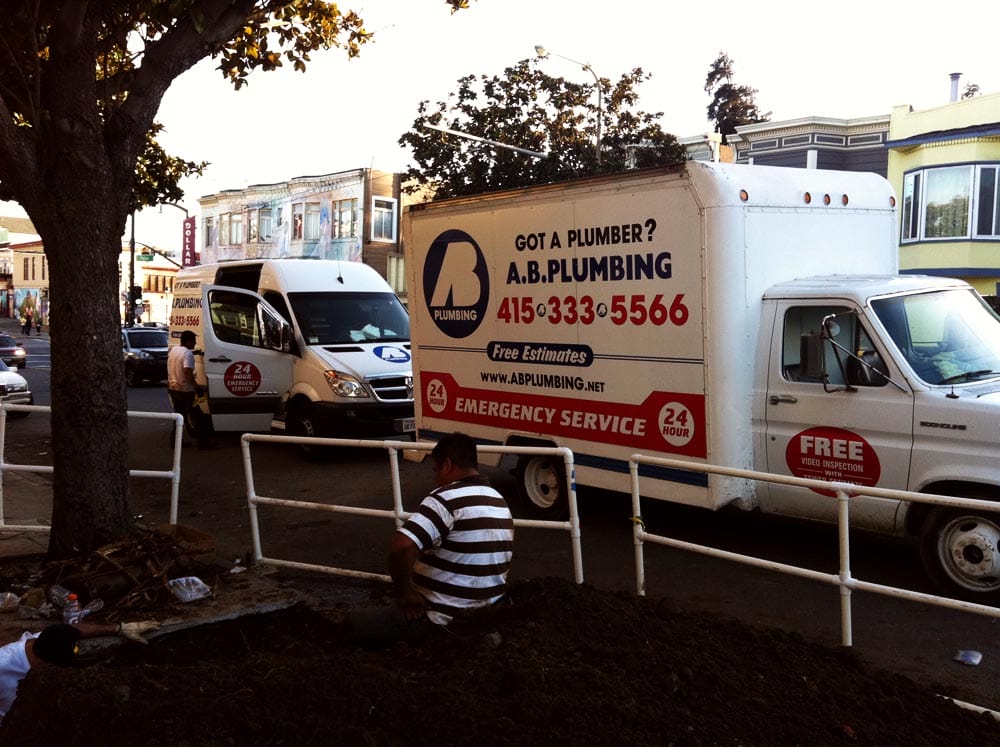 A plumber works near a tree on a sidewalk. Two plumbing services trucks are parked nearby, displaying contact information and offering free estimates and 24-hour emergency service.