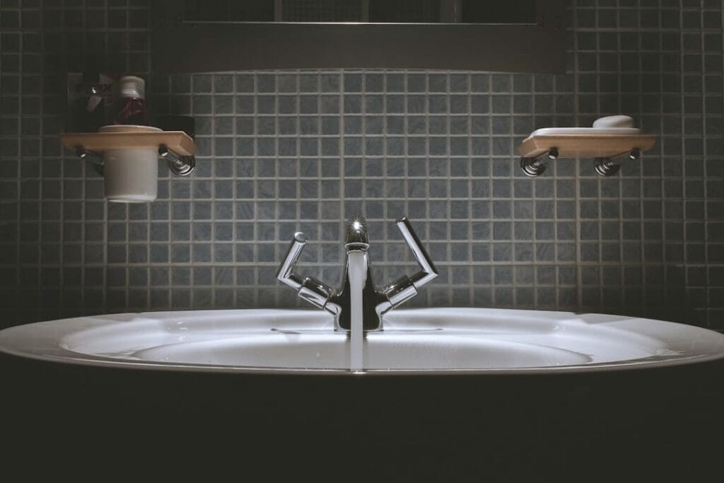 A bathroom sink with a modern faucet is centered under a tiled wall. Small shelves holding soap and a cup are mounted on either side above the sink.